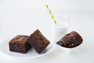 Slice of delicious homemade chocolate brownie served on white plate. Served with glass of milk with green white stripe straw. Taken on a white background.
