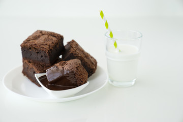 Slice of delicious homemade chocolate brownie served on white plate. Served with glass of milk with green white stripe straw. Taken on a white background.