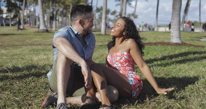Young stylish black woman with cheerful man sitting on lawn in park and chatting with laugh having fun in sunlight. 
