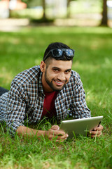 Naklejka na ściany i meble Smiling Indian man lying on green lawn and posing for photography while surfing Internet on modern digital tablet, portrait shot
