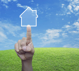 Hand pressing house icon with copy space over green grass field with blue sky, Real estate concept