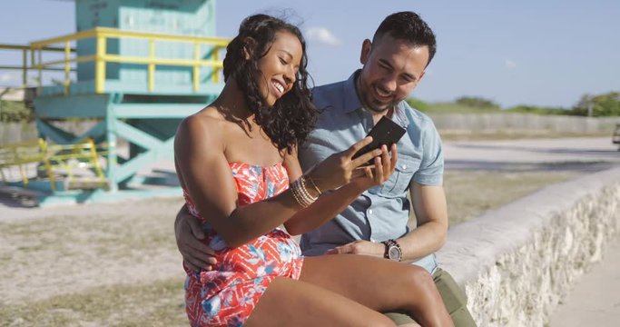 Beautiful multiracial man and woman embracing on waterfront in bright sunlight and taking selfie with smartphone. 