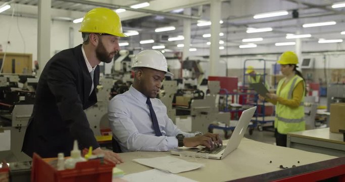 Manager and foreman working on a laptop in a factory discussing business.