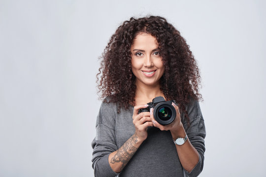Smiling woman photographer with professional photo camera, front view portrait over grey background