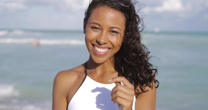 Headshot of bright African-American woman with curly hair wearing white bikini and laughing at camera on shoreline of ocean. 