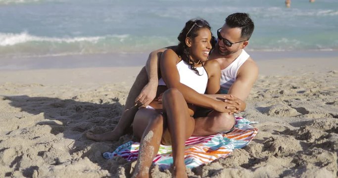 Charming black girl in white bikini embracing with handsome man on towel chilling on beach in sunlight with ocean on background. 