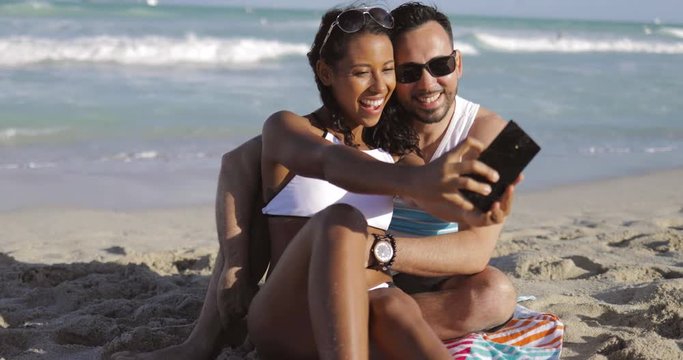 Content black girl with man sitting on sandy beach and using camera of smartphone and taking selfie for memory. 