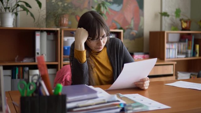 The young female teacher in glasses check independent tests sitting at a table in the elementary school classroom.