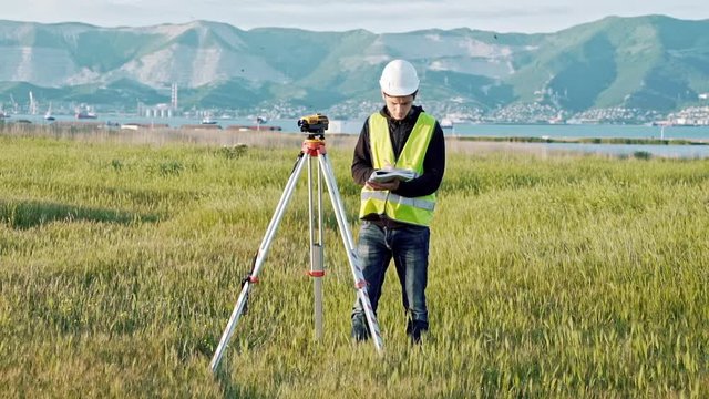 surveyor man in green work clothes and helmet adjusts the equipment, produces calculations and writes to the notebook on the construction site. The concept of landscape design, geodesy