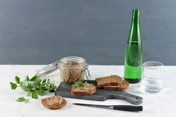 On a wooden board slices of black bread with liver pate. Next to the pot with pâté. In the frame, a knife, parsley, a bottle of water, a glass. Light surface. Close-up.