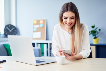 Beautiful smiling woman using phone while working on laptop in home office