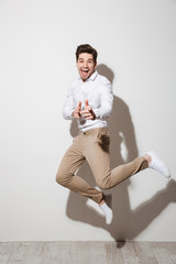 Full length photo of handsome man 30s in stylish clothing and sneakers jumping and pointing index fingers on camera isolated over white background with shadow