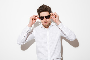 Fashion portrait of fancy man model dressed in shirt touching sunglasses and looking on camera with serious gaze, over white background with shadow