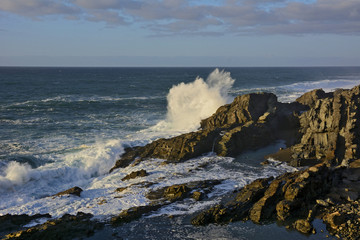 Felsige Meeresküste mit einer Welle die sich an einem Felsen hoch aufspritzend bricht.