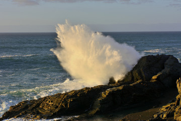 Meeresküste mit einer Welle die sich an einem Felsen hoch aufspritzend bricht.