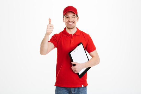 Positive deliveryman in red t-shirt and cap smiling and showing ok symbol while holding clipboard, isolated over white background