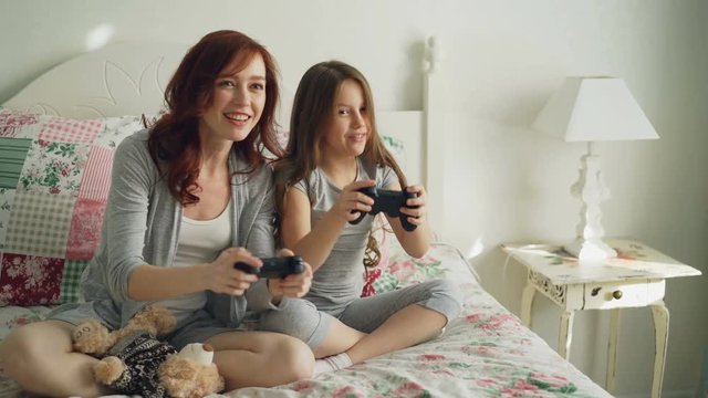 Funny laughing girl with happy young mother have fun while playing computer console games on TV sitting on bed at home in the morning in cozy bedroom. Daughter embracing mom after winning