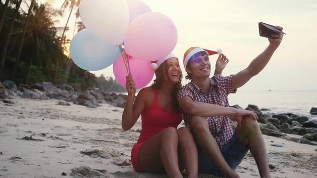 Happy funny couple sit on the beach with multicolored balloons on Christmas travel holidays taking selfie picture with smartphone wearing santa hat during beautiful sunset in slow motion. 1920x1080