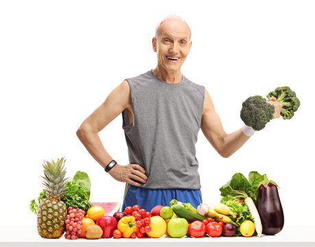Elderly man holding a broccoli dumbbell behind a table with fruit and vegetables