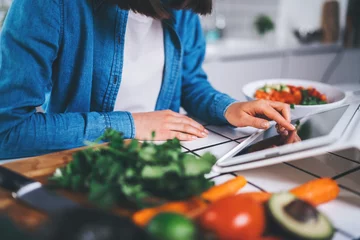 Fotobehang Closeup of female cook using digital tablet device for searching healthy meal recipes, young woman sitting in the kitchen preparing vegetarian food or salad, eating raw food © iana_kolesnikova