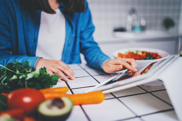 Closeup of female food blogger using digital tablet device looking for healthy food recipes, focus on a hands, cropped image of young woman sitting in modern kitchen using portable computer