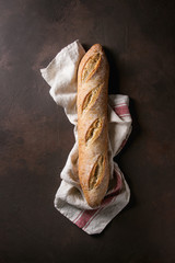 Loaf of fresh baked artisan baguette bread on linen cloth over dark brown texture background. Top view, copy space.