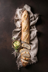 Loaf of sliced fresh baked artisan baguette bread on linen cloth with butter and herbsover dark...