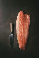 Whole raw uncooked salmon fillet with chef's knife over dark brown texture background. Top view,...