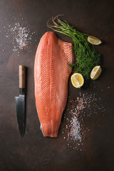 Whole raw uncooked salmon fillet server with green dill, lemon, salt, pepper, chef's knife over...
