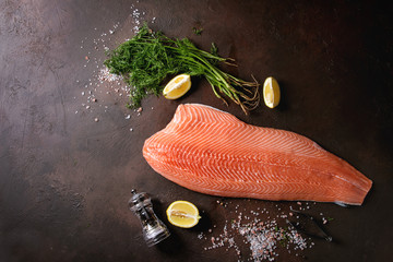 Whole raw uncooked salmon fillet server with green dill, lemon, salt, pepper, chef's knife over...