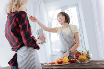 Try it. Joyful nice happy woman standing at the table and smiling while feeding her girlfriend