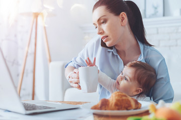 Very busy. Serious attractive good looking woman holding a cup of tea and talking on the phone while looking after her child