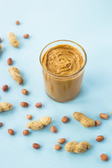 A glass jar with peanut butter paste among the peanuts in shell and without on a blue background....