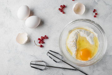 Top view photo of a broken egg on flour in a glass plate. Mixer fits on a white surface. Eggs and currant on a table. Ingredients for healthy rustic breakfast.