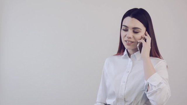 Young business woman talking on the phone in the office. Girl in white shirt speaks on smarfona indoors. Pleasant, quiet phone conversation. The girl cute talks, smiles, answers a phone call. 