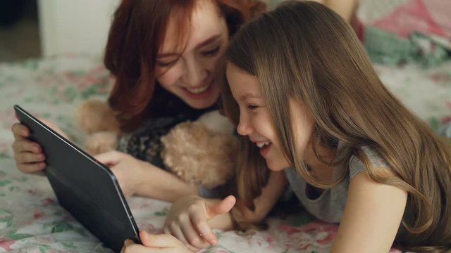 Close-up of attractive young mother and her cute daughter in pajamas laughing and looking in digital tablet while lying on bed at home in the morning