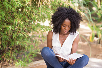 Focused black young woman reading book in city garden. International student preparing for classes...