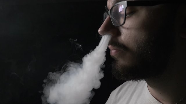 Young teenager student boy with beard moustache and glasses vaping electronic cigarette blowing smoke on his nose and then on his mouth in slow motion