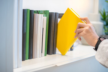 male hand choosing and picking white book on white bookshelf in public library, education research...