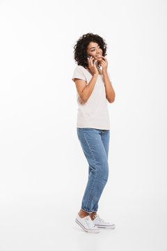 Full length picture of content american woman with afro hairstyle wearing t-shirt and jeans speaking on cell phone with pleasure, isolated over white background