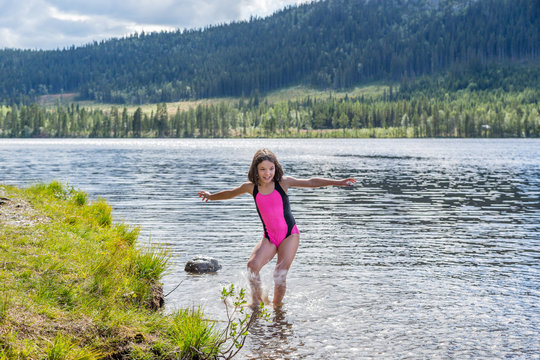 Cute little brown haired girl in swimsuit spashing water in a mountain lake.