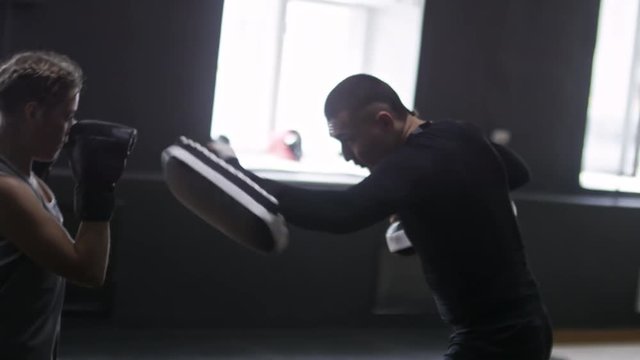 Sporty young woman in boxing gloves kicking and punching her male instructor, who is using boxing kick pads for protection