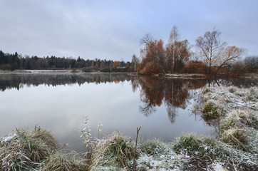Cloudy autumn landscape with birches growing on the bank of pond.First snow and hoarfrost in november.Overcast sky.Moscow region,Russia. 