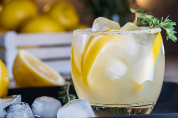 Lemon cocktail with thyme and ice on dark rustic background, close-up. Refreshing alcoholic yellow cocktail drink.