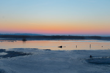 the sun rises red over marshlands and swamps and a wooden bridge with a railing and birds resting on the water and among the reeds