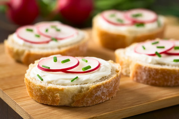 Bread slices with cream cheese, radish slices and chives on wooden board, photographed with natural light (Selective Focus, Focus on the front of the radish slices on the first bread)