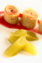 Fresh homemade rhubarb compote with crepe rolls filled with strawberry jam in the back, photographed with natural light (Selective Focus, Focus on the front of the first rhubarb piece)