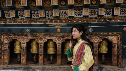 Bhutanese young woman wearing a kira (national dress) standing in front of prayer wheels in...