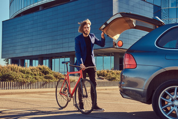 A bearded fashionable bearded male with long hair takes out a bicycle from the car's trunk.