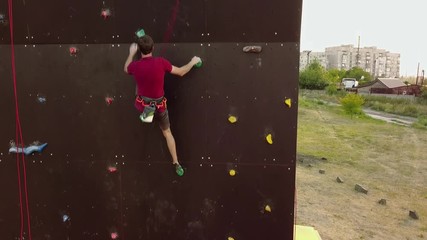 Aerial of climber runs the speed rock climbing track on artificial wall outdoors. Young fast athlete climbs the cliff at competitions. Man on belaying harness performs at bouldering championship.